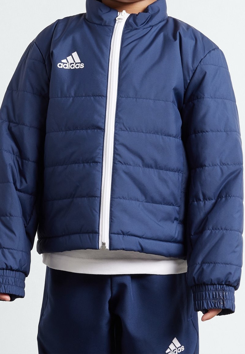 Námorně modrá péřová bunda, prošívaný design, bílý zip, vysoký límec, pružné manžety, s kontrastním bílým logem Adidas na hrudi.