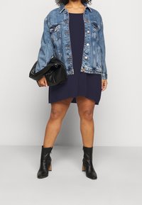 Jeansjack met een lichte wassing, knoopsluiting, twee voorzakken, over een donker marineblauwe jurk. Zwarte, gestructureerde handtas en hoge enkellaarsjes.