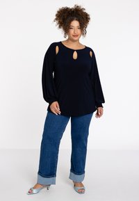 Marineblauw langmouwtop met sleutelgatdetails, elastische manchetten, gecombineerd met blauwe denim wijde broek en lichtblauwe haksandalen.