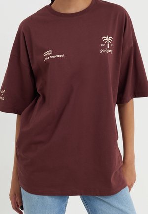 Persoon draagt een losse kastanjebruine T-shirt met witte tekst en een palmboom-design, gecombineerd met lichtblauwe spijkerbroek.