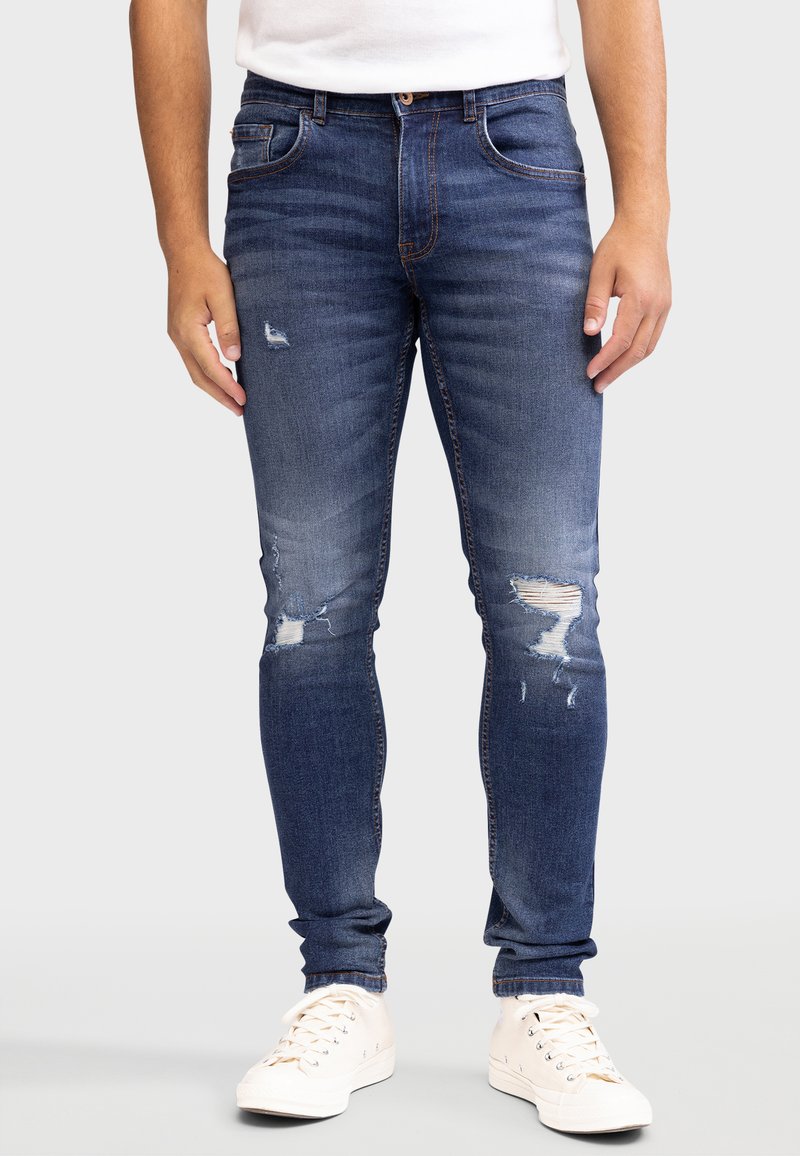 Vaqueros ajustados de color azul oscuro con ligero desgastado y parches de rodilla deshilachados. Diseño clásico de cinco bolsillos y costuras en contraste. Llevados con zapatillas blancas.