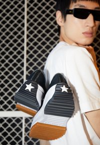 Baskets noires et blanches avec semelles en caoutchouc texturées et une semelle extérieure marron, présentant un accent étoile blanche sur le talon, tenues contre un fond en maille.