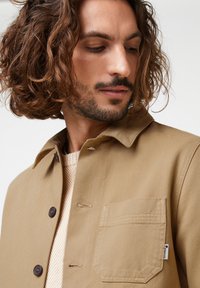 Lichtbruine katoenen jas met een kraag, bruine knopen en een borstzak. Getextureerde stof met een strak ontwerp en minimale details.