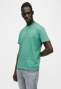 Πράσινο βαμβακερό t-shirt με στρογγυλή λαιμόκοψη, με χρυσό λογότυπο "TOMMY JEANS" στο στήθος. Κοντά μανίκια και άνετη εφαρμογή.