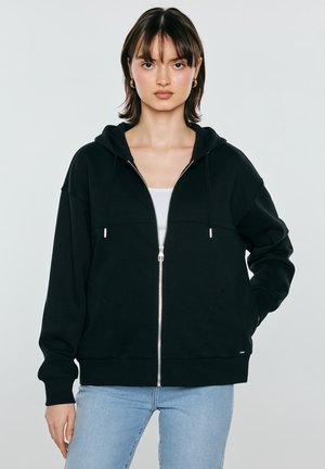 Młoda kobieta o krótkich ciemnych włosach, nosząca złote kolczyki-koła, czarną bluzę z zamkiem na suwak, biały top i jasnoniebieskie dżinsy na neutralnym tle.