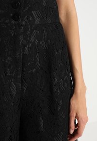 Černé krajkové kalhoty s propracovanými květinovými vzory a texturou, se dvěma černými knoflíky v pase. Ruka je umístěna vedle.