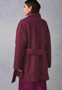 Burgundy villakangastakki vyötäröltä säädettävällä vyöllä, ylisuuri malli ja lovettu kaulus. Teksturoitu kangas minimalistisella muotoilulla, pitkähihaisilla hihoilla.
