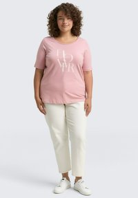Roze t-shirt met korte mouwen en een crèmekleurige "FLOWER" graphic, gecombineerd met witte rechte broek en witte sneakers op een neutrale achtergrond.