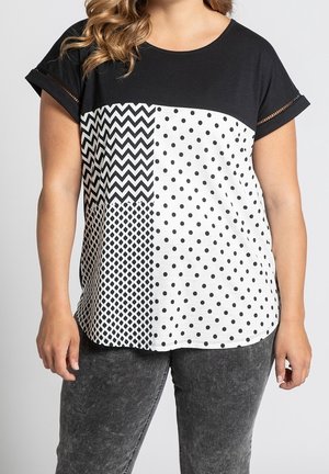 Frau trägt ein kurzärmeliges schwarz-weißes Oberteil mit Zickzack-, Polka-Dot- und Diamantmustern, kombiniert mit dunkelgrauen Jeans.