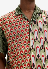 Kortärmad mönstrad skjorta med grön bas, med röda, svarta och blå mönster; geometrisk krage; knäppning framtill.