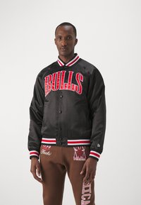 Svart satin bomberjacka med röd "BULLS"-text, ribbad krage och muddar med röda och vita ränder, stängd med tryckknappar.