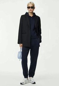 Μαύρη σακάκι πάνω από ναυτική κουκούλα, συνδυασμένο με ναυτικά joggers και λευκά αθλητικά παπούτσια. Τα αξεσουάρ περιλαμβάνουν μια ανοιχτό μπλε τσάντα και σκούρα γυαλιά ηλίου.