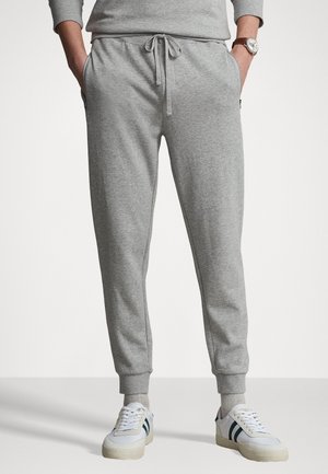Person trägt hellgraue Jogginghose mit Kordelzug, gesäumte Knöchel, Hände in den Taschen, hellgraue Socken und weiße Sneakers mit dunklen Streifen.