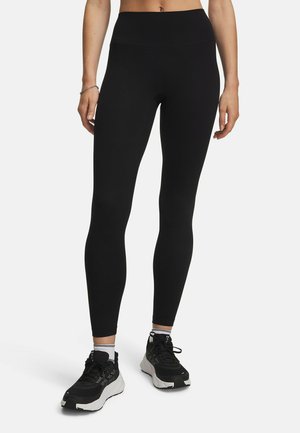 Vrouw draagt zwarte leggings met hoge taille en zwart-witte sportschoenen, staat met ontspannen armen langs haar zij op een witte achtergrond.
