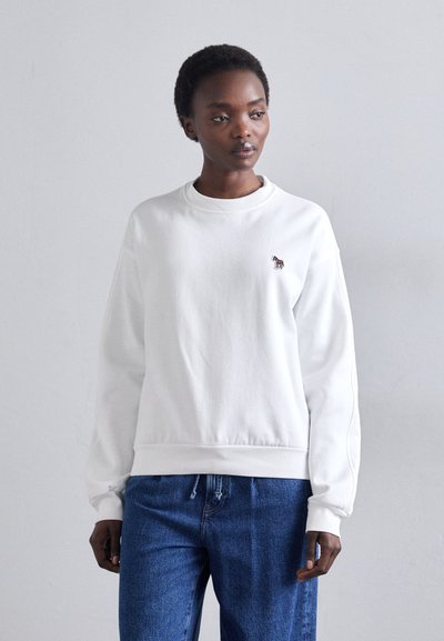 Sweatshirt blanc avec un col rond, des poignets et un ourlet côtelés, présentant une petite broderie de zèbre multicolore sur la poitrine gauche. Jean bleu.