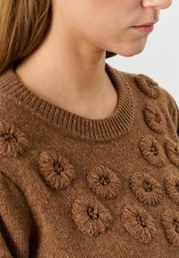 Brauner Strickpullover mit rundem Ausschnitt, der strukturierte Blumenmuster aus kontrastierendem Garn auf der Vorderseite aufweist. Weiches, gemütliches Material.