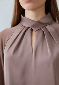 Schlichte Bluse aus staubrosafarbenem Stoff mit hohem Kragen, plissierten Details und einer Schlitzöffnung am Kragen. Glatte Textur, elegantes Design.