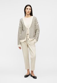 Kvinde stående iført mønstret beige cardigan, hvid top, beige bukser med høj talje og knapper samt sorte flade sko mod hvid baggrund.