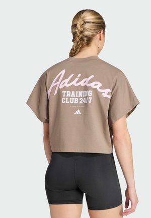 Donna con capelli biondi intrecciati che indossa una maglietta corta marrone Adidas con la scritta "Training Club 24/7" e pantaloncini sportivi neri, con la schiena rivolta verso la fotocamera.