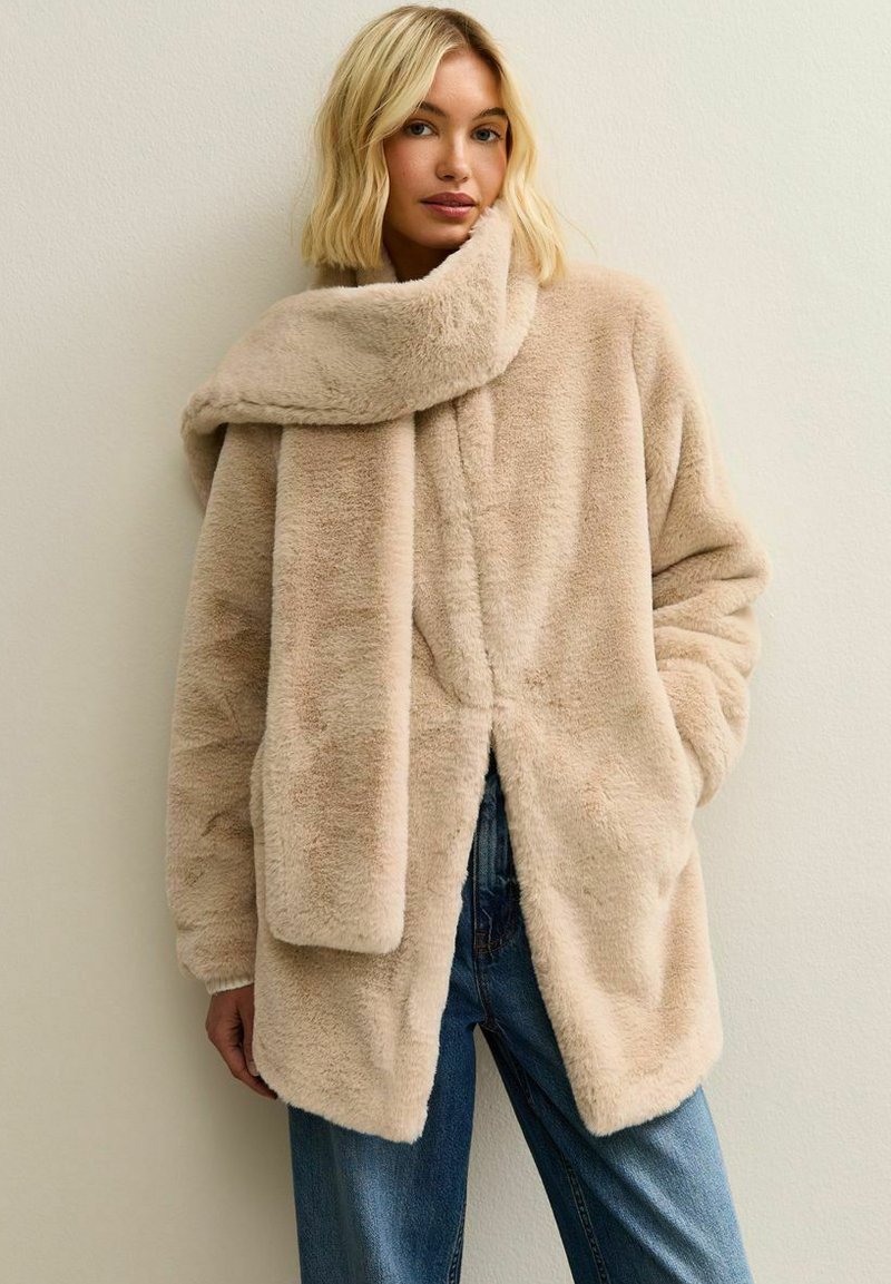 Manteau en fausse fourrure beige avec un large col, des manches longues et une coupe décontractée. Présente une fente à l'avant et une texture légère. Porté par-dessus un jean en denim bleu.