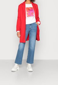 Un long manteau rouge sur un t-shirt graphique blanc avec un design floral, associé à des jeans évasés bleu clair et des bottines blanches.