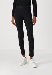 Zwarte leggings van rekbaar materiaal, aansluitend ontwerp, met een getextureerde tailleband met branding, gecombineerd met witte sneakers.