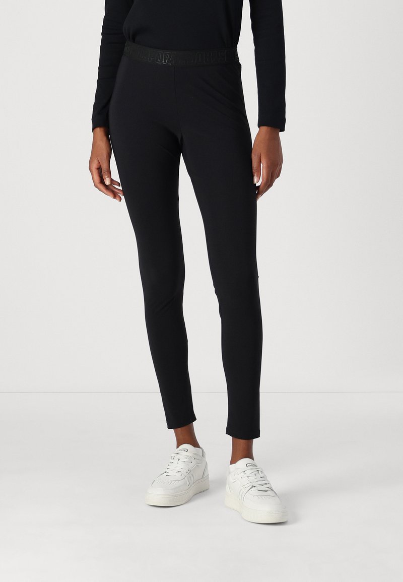 Zwarte leggings van rekbaar materiaal, aansluitend ontwerp, met een getextureerde tailleband met branding, gecombineerd met witte sneakers.