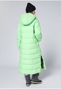 Langer, wattierter grüner Daunenmantel mit Kapuze; verfügt über ein verstecktes Leopardenmuster-Futter und seitlichen Schlitz. Kombiniert mit grauen Winterstiefeln.