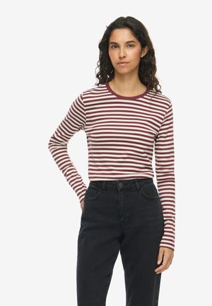 Ung kvinde med krøllet hår iført en langærmet trøje med bordeaux- og hvide striber og sorte højtaljede jeans, står med den ene hånd på hoften.