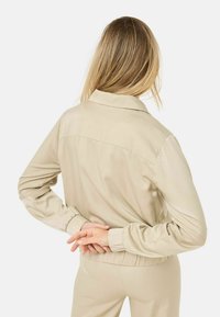 Giacca sintetica beige con colletto, polsini elasticizzati e vita arricciata. Texture liscia con design semplice.
