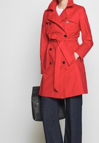 Femme portant un trench-coat rouge ceinturé par-dessus un jean foncé, tenant un sac fourre-tout en cuir noir, debout devant un fond uni.