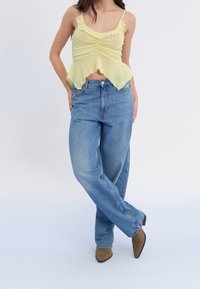 Top sin mangas de color amarillo claro con bordes acampanados, combinado con jeans de pierna recta de tiro alto en azul claro y botas de tobillo de punta marrón.