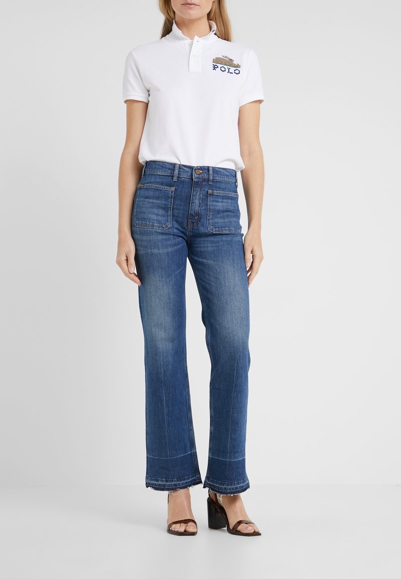 Vit pikétröja med krage och broderad "POLO"-logotyp, tillsammans med mörkblå vida jeans med framfickor och fransiga nederkanter.