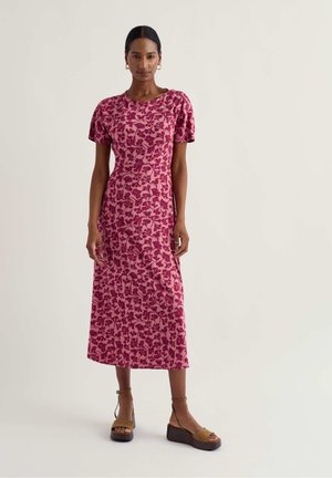 Nő áll, lila virágos midi ruhát visel, puffos ujjakkal és barna platform szandállal, egyszerű világos háttér előtt.