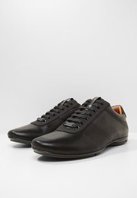 Chaussures en cuir noir avec un design perforé, une texture lisse, un bout arrondi et des lacets noirs. Présentent un logo sur la langue et le côté.