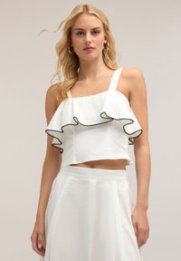 Top bianco corto con orlo a volant fluido, caratterizzato da rifiniture nere. Ha spalline larghe e un design aderente al busto. Tessuto dalla consistenza liscia.