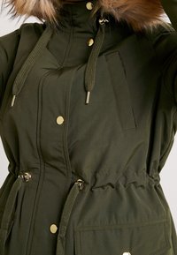 Olivově zelená parka s kapucí podšitou kožešinou, zdobená zlatými knoflíky, stahovacími šňůrkami, elastickým pasem a přední kapsou.