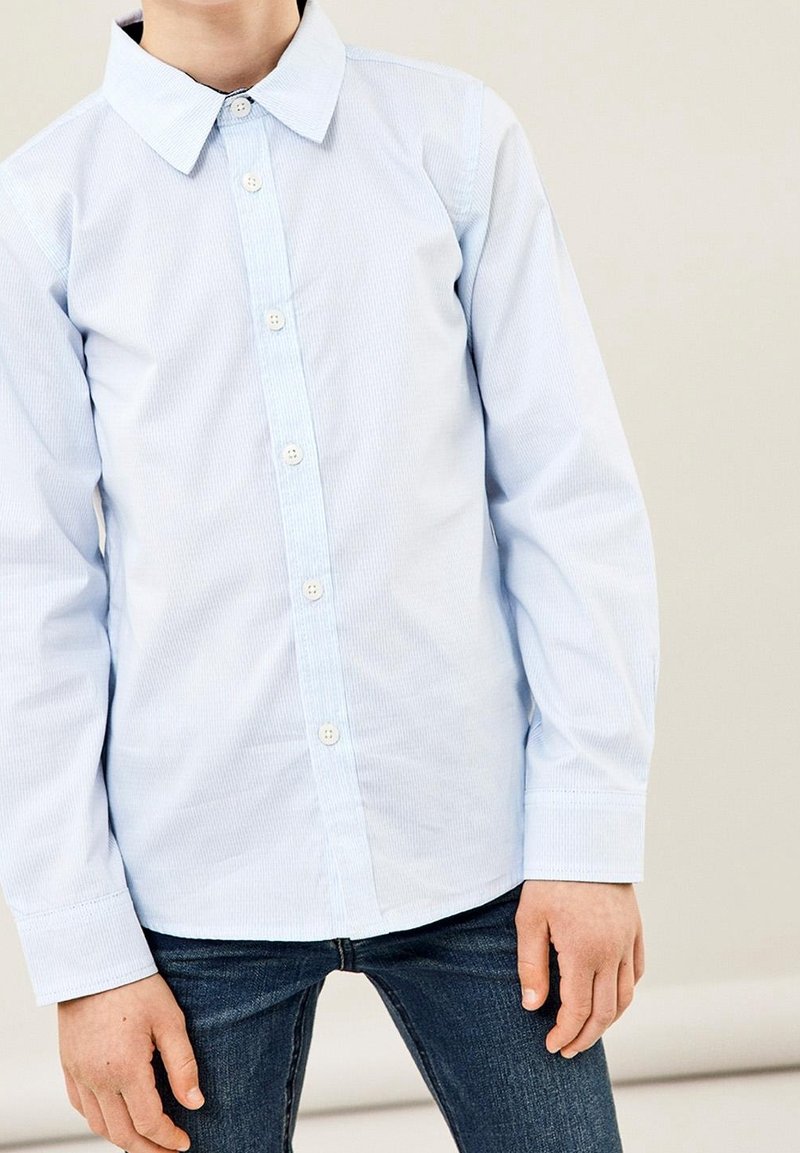 Enfant portant une chemise boutonnée bleu clair et un jean bleu foncé, debout devant un fond clair uni.