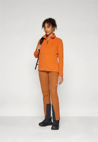 Oranž värvi fliisjakk esiküljel lukuga, kombineeritud pruunide pükstega, mille varrukate ääris on mustad detailid. Model kannab musti matkasaabastega jalanõusid.