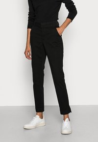 Pantalons noirs fuselés en tissu mélangé coton, avec poches latérales, texture lisse et fentes à l'ourlet, associés à des baskets blanches.