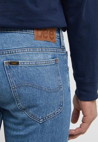 Niebieskie dżinsy z denimu z skórzaną naszywką marki Lee na tylnej części w pasie, charakteryzujące się klasycznym designem pięciu kieszeni i kontrastowym przeszyciem.