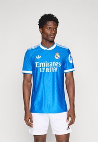Blue soccer jersey with white accents, featuring "Emirates Fly Better" text, the Real Madrid logo, and embroidered stripes on the sleeves.
