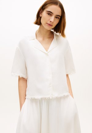 Ung kvinde med kort brunt hår iført en løs hvid skjorte med knapper og frynser samt matchende hvid nederdel, hænderne i lommerne.