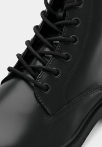 Stivale in pelle nera con finitura liscia, dotato di lacci neri, occhielli in metallo e punta arrotondata. Le cuciture sono visibili lungo la cucitura.
