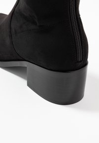 Botín de ante negro hasta el tobillo con un bajo tacón cuadrado, que presenta un cierre lateral y una textura suave. El diseño es elegante con una estética minimalista.