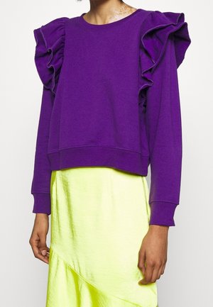 Personne portant un haut violet à manches longues avec des épaules volantées et une jupe jaune brillante, montrée du cou aux hanches.