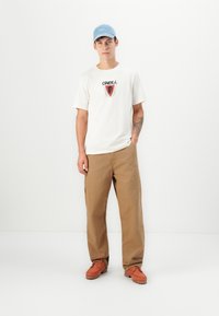 White cotton t-shirt with black and red graphic, tan wide-leg pants, and brown lace-up shoes. Light blue cap completes the look.