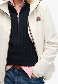 Biała wodoodporna kurtka z odpinanym kapturem, granatowy sweter ze ściągaczami pod spodem, różowa koszula z guzikami, oraz niebieskie dżinsy. Posiada naszywkę z logo.