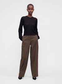 Femme debout, les mains dans les poches, portant un haut noir à manches longues, un pantalon large marron et des chaussures noires pointues sur un fond clair uni.