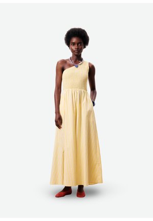 Femme portant une robe longue jaune à une épaule, chaussures plates rouges, collier de perles bleu, debout avec une main dans la poche.